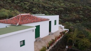 Un edificio blanco con tejados rojos y contraventanas verdes, rodeado de abundante vegetación y plantas en macetas.