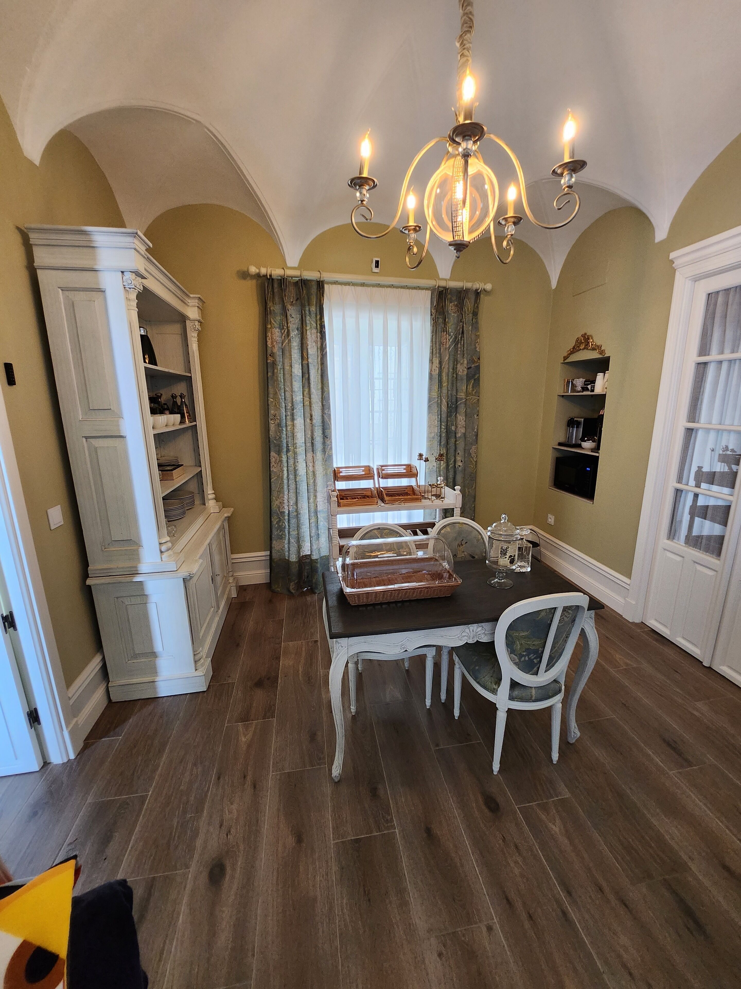 A dining area with a chandelier, white chairs, a table with a glass cover, and a buffet.