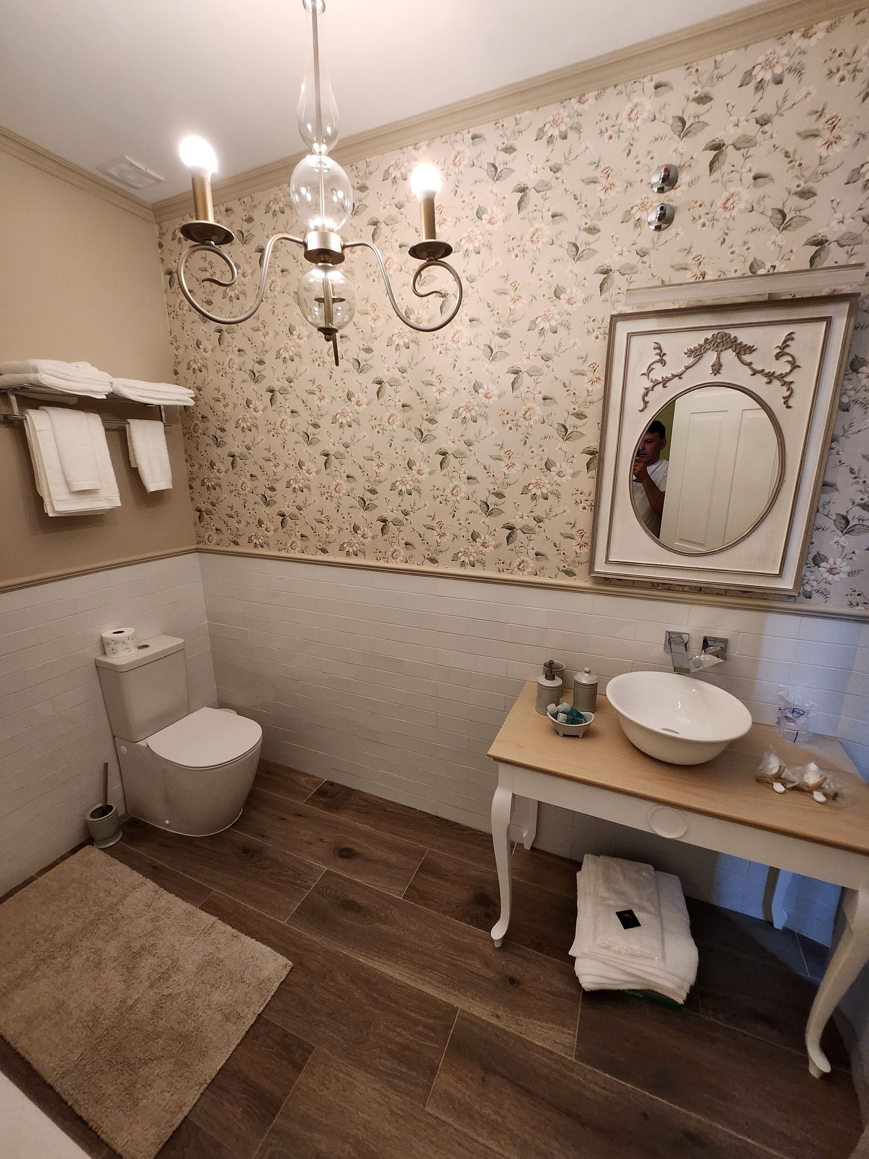 Un baño con inodoro, lavamanos y espejo. Hay toallas en un estante, y un candelabro cuelga del techo.