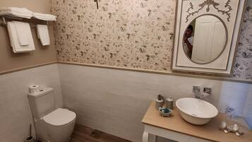Un baño con inodoro, lavamanos y espejo. Hay toallas en un estante, y un candelabro cuelga del techo.