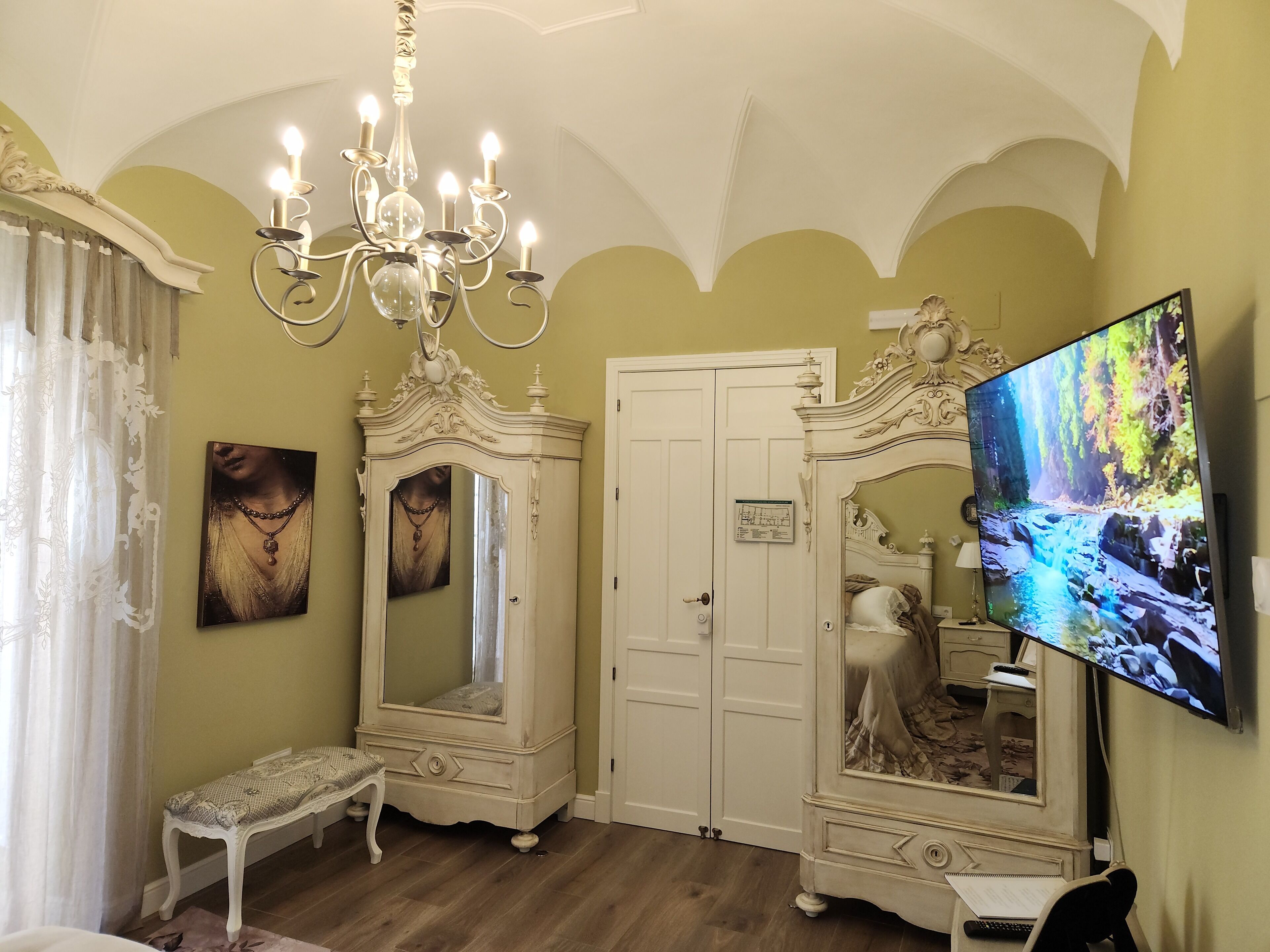 Una habitación con candelabro, un espejo grande, televisor y un banco.