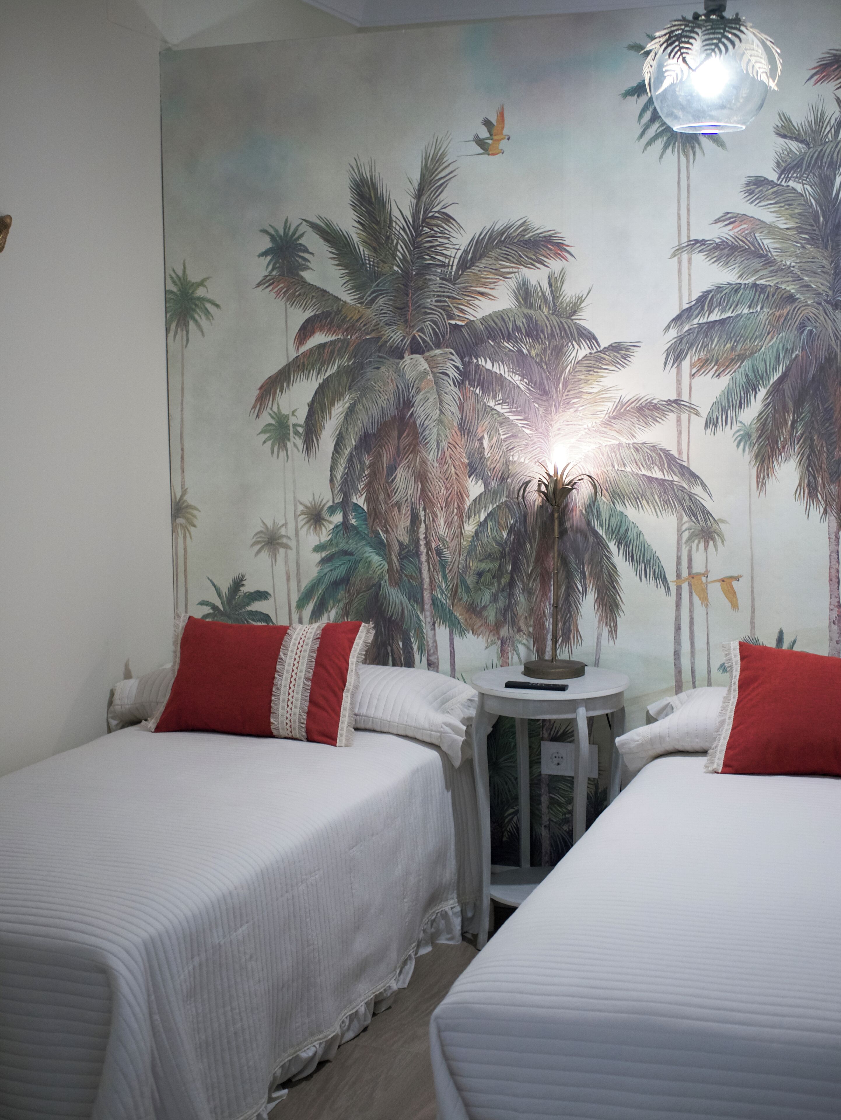 Una habitación con dos camas, cada una con ropa blanca y almohadas rojas, y un mural de palmeras en la pared.