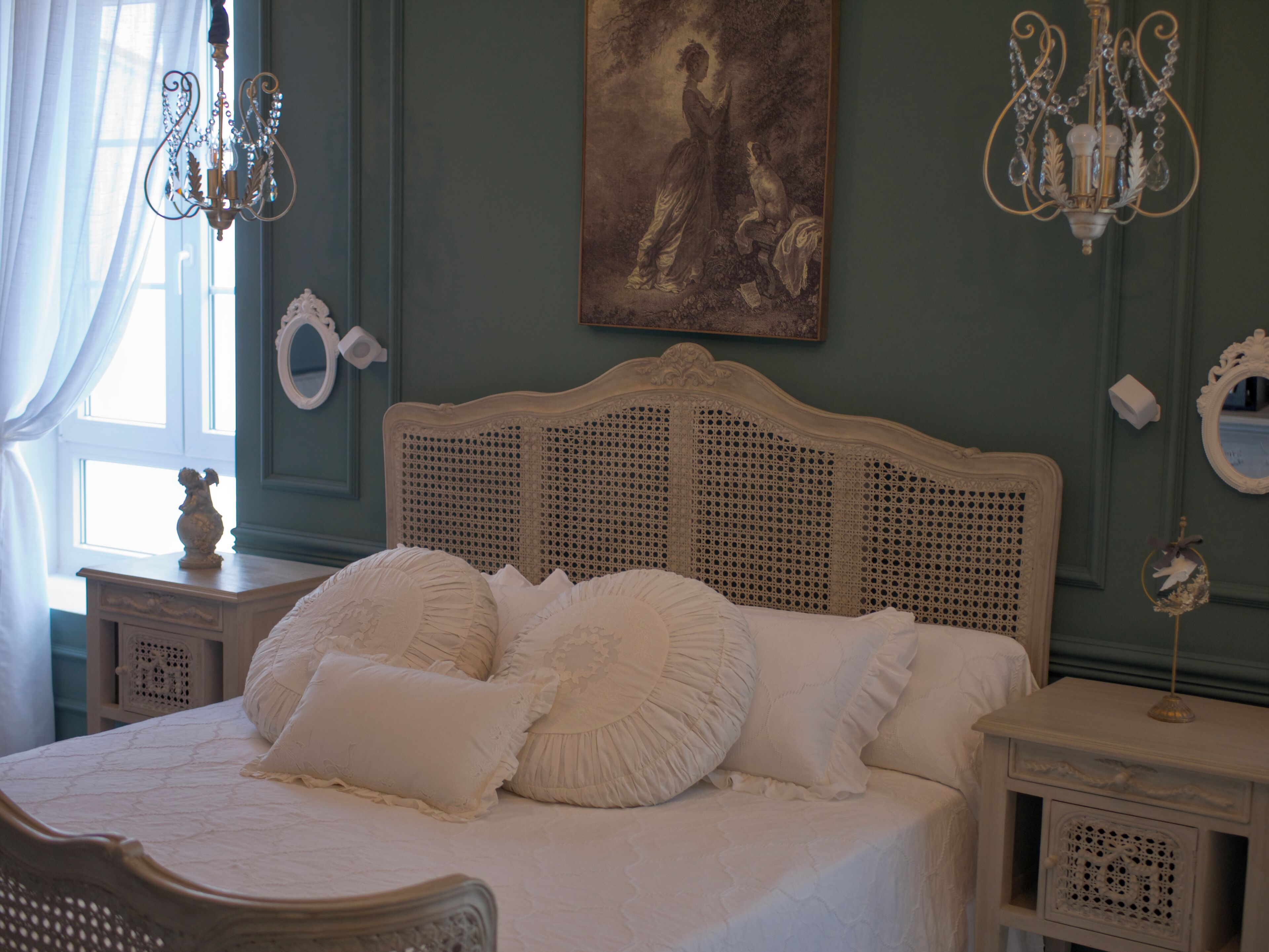 A bedroom with a bed, two nightstands, a chandelier, a painting, and a window with curtains.