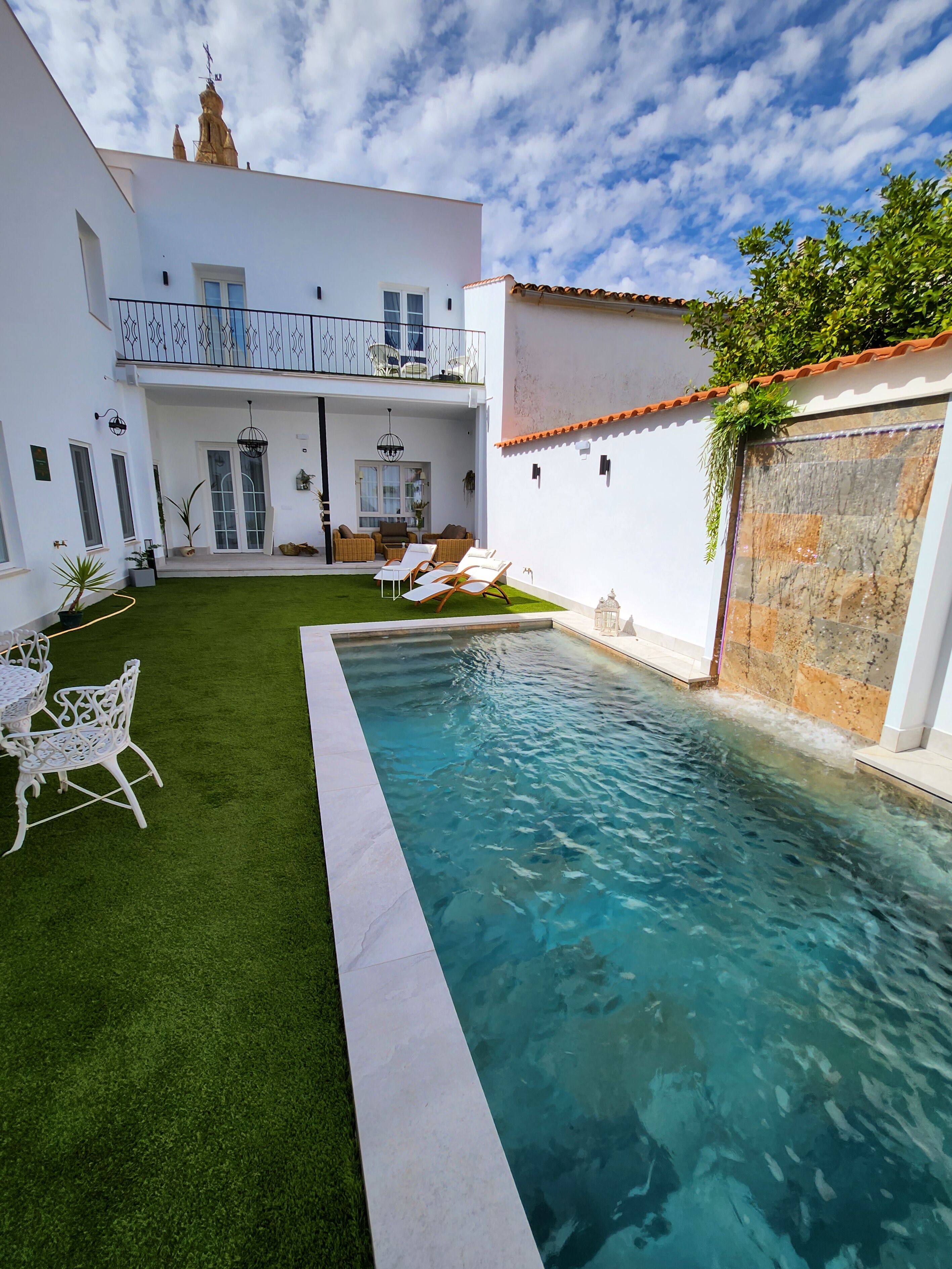 Una piscina con borde blanco, rodeada por un césped cuidado y mobiliario exterior blanco.