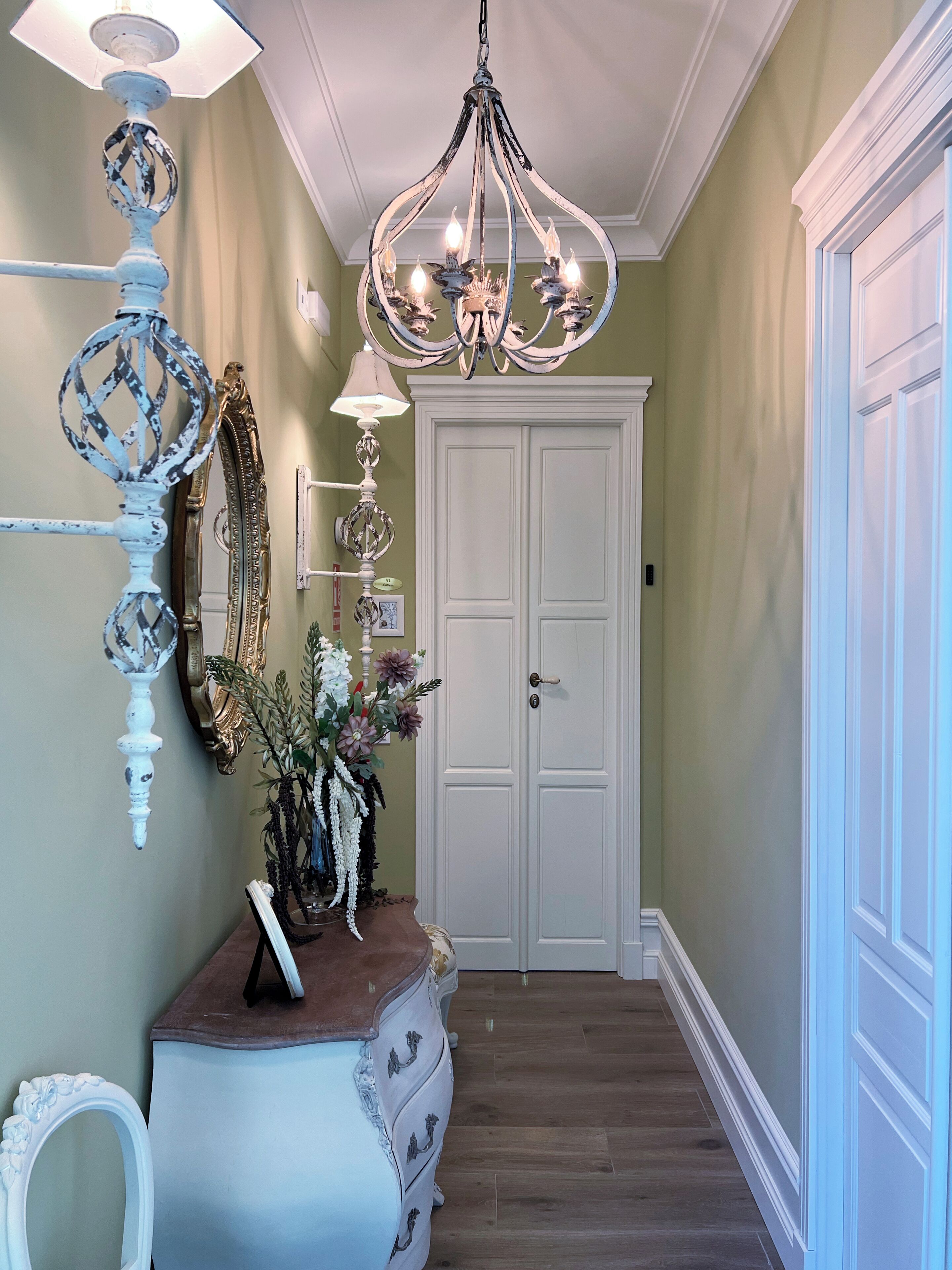 Un couloir avec une porte blanche, un lustre, un miroir et une commode sur laquelle est posé un vase de fleurs.