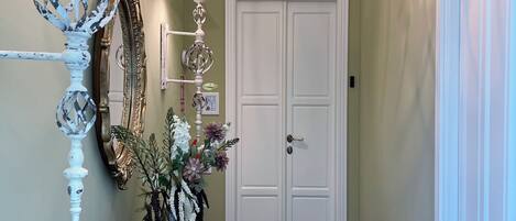 Un couloir avec une porte blanche, un lustre, un miroir et une commode sur laquelle est posé un vase de fleurs.