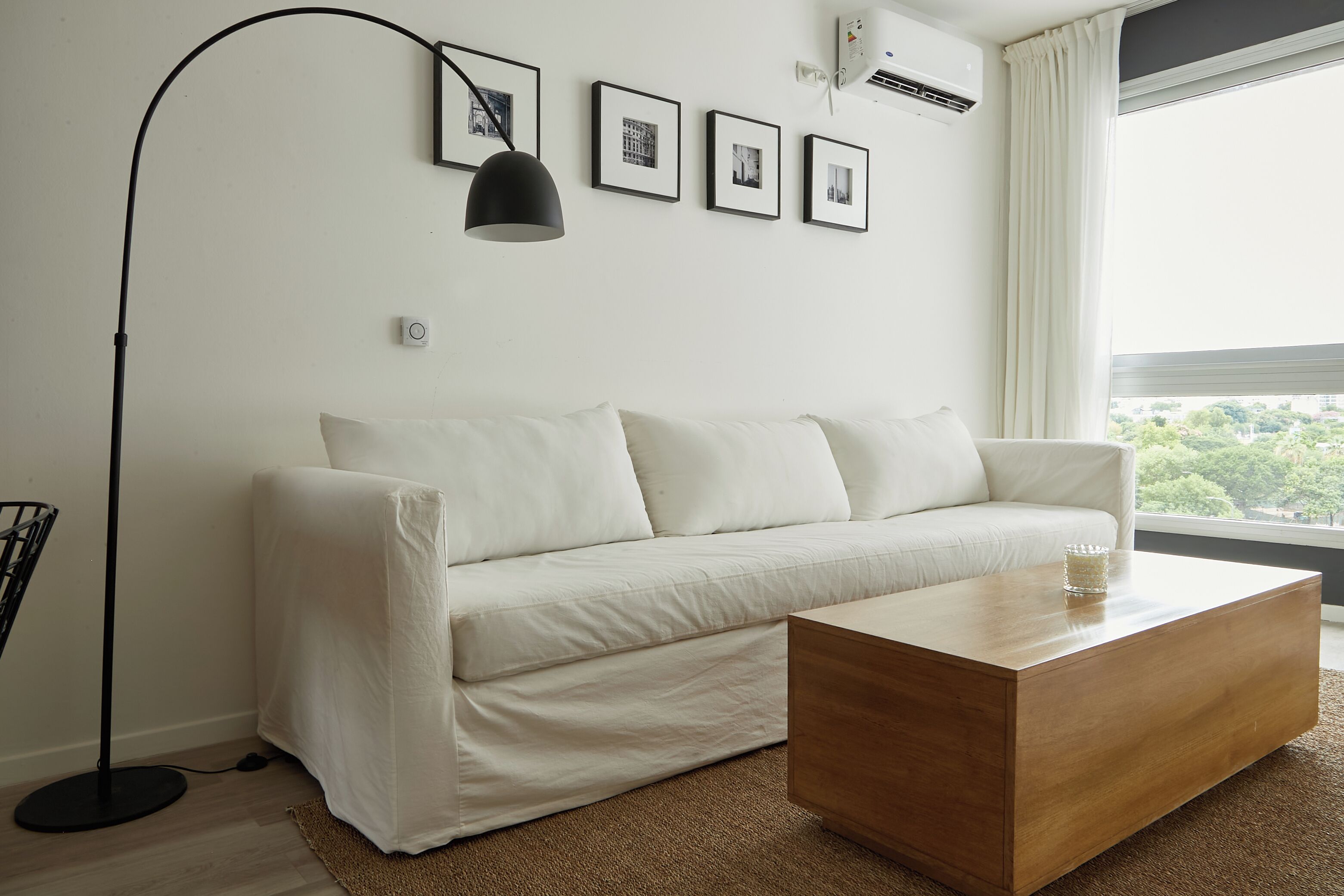 Una sala moderna con un sofá blanco, una mesa de centro de madera y una lámpara de pie. Se observan cuadros enmarcados en la pared y un equipo de aire acondicionado.