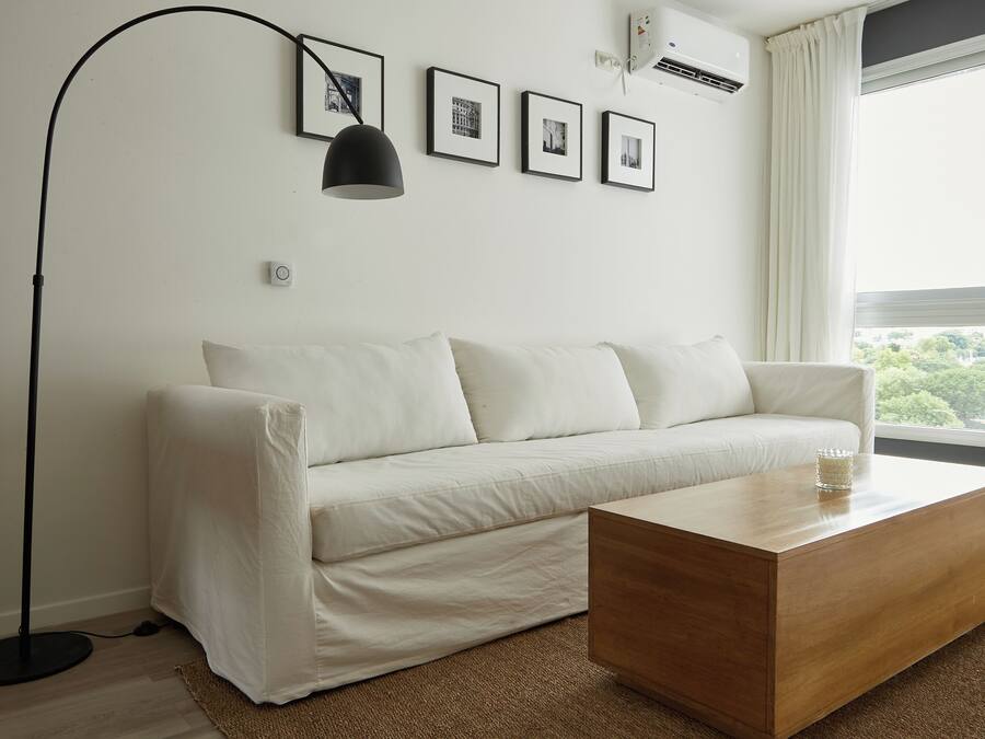 Una sala moderna con un sofá blanco, una mesa de centro de madera y una lámpara de pie. Se observan cuadros enmarcados en la pared y un equipo de aire acondicionado.