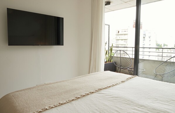 Una camera da letto con un letto, una TV a parete e un balcone con sedie e un vaso con una pianta.