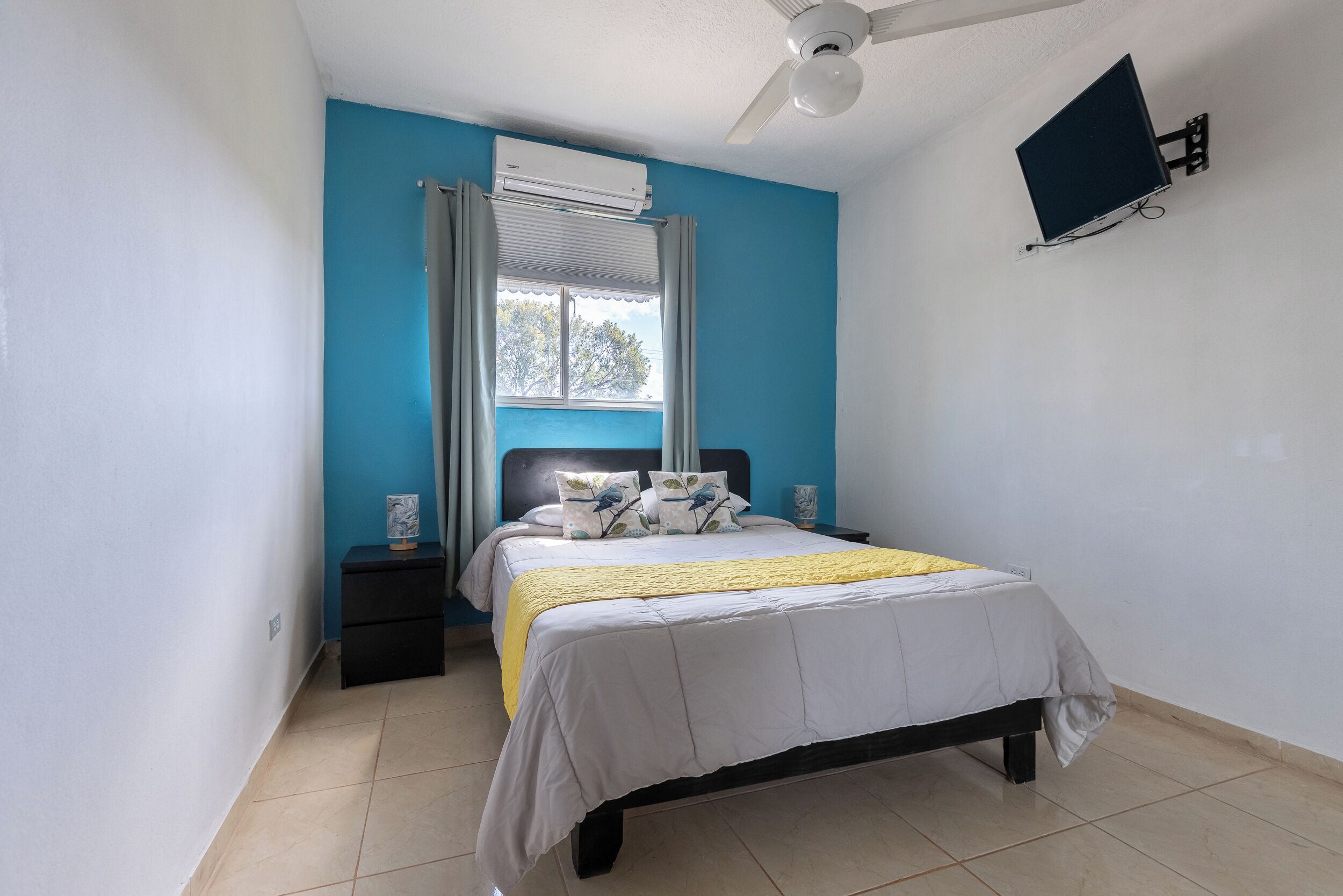 Een slaapkamer met een bed, nachtkastje, een aan de muur bevestigde televisie en een airconditioner.