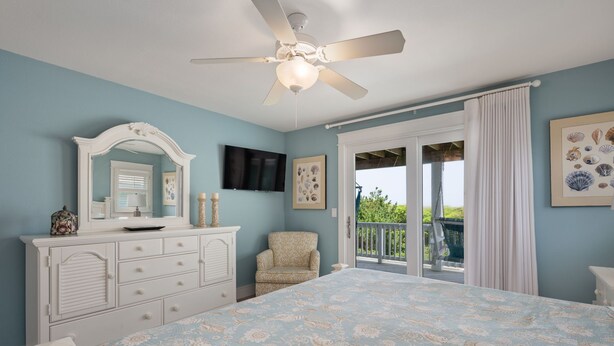 A bedroom with a bed, dresser, chair, ceiling fan, and a balcony with curtains.
