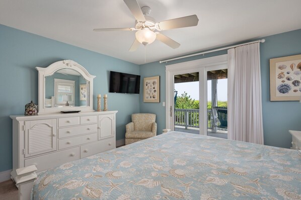 A bedroom with a bed, dresser, chair, ceiling fan, and a balcony with curtains.