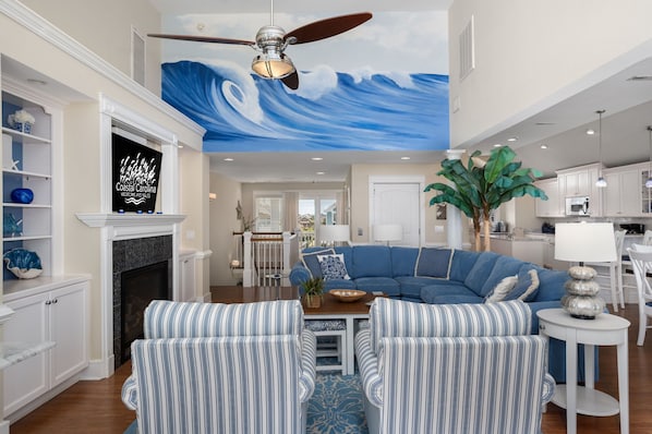 A living room with a blue wave mural, a ceiling fan, a fireplace, and a dining area with a white table and chairs.