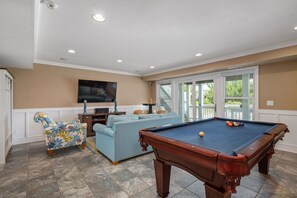 A pool table, a sofa, and a television in a room with beige walls and a tiled floor.
