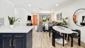 Een moderne keuken met donkere kasten en een wit aanrecht, een eethoek met een ronde tafel en stoelen, en een woongedeelte met een bank en salontafel.
