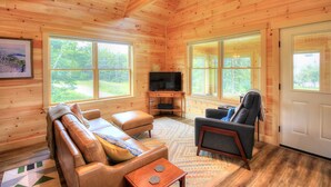Ein gemütliches Blockhauss-Interieur mit einem Ledersofa, einem Relaxsessel, einem Fernseher und großen Fenstern mit Blick ins Freie.
