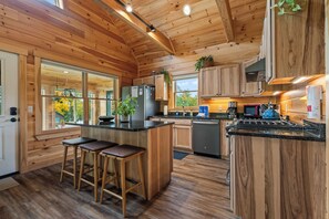 A modern kitchen with wooden cabinetry, a central island with stools, a refrigerator, and a coffee machine. - Niboban Cabin 17 - shared waterfront (Rangeley)