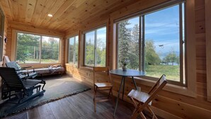 Ein gemütliches Blockhaus-Interieur mit Holzwänden und -böden, großen Fenstern mit Blick ins Freie, einer Sitzecke mit einer Bank und Stühlen sowie einem kleinen Tisch mit einer Vase.