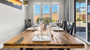 A dining area with a long wooden table set for a meal, large windows with blinds, and a cityscape painting on the wall. - Escondido 1149, Desert Bloom Retreat|Grill, Smart TV, Xbox 360, 2 Laundry Rooms, SLP 14 (Washington)