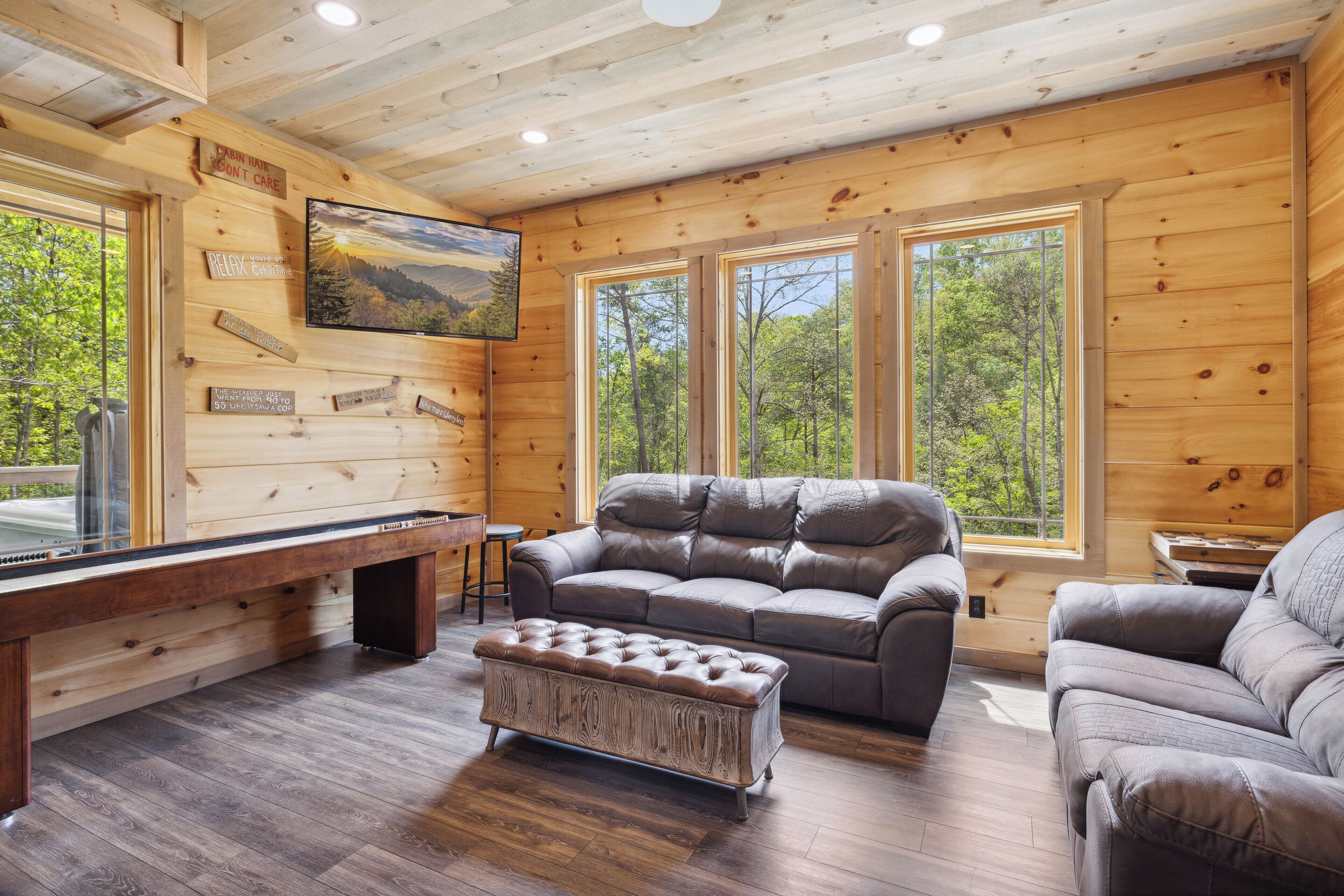 Un intérieur de chalet chaleureux avec une table de billard, des canapés en cuir et de grandes fenêtres donnant sur la forêt.