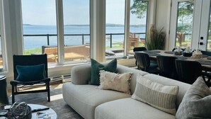 A spacious living room with a large window overlooking a body of water. There is a beige sofa with multiple cushions, a round coffee table, and a dining area with chairs and a table. - Dreamy Oceanfront Escape Hot tub & Sauna (South Shore)