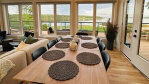 A spacious dining area with a long wooden table, multiple chairs, and large windows offering a view of a lake and greenery. - Dreamy Oceanfront Escape Hot tub & Sauna (South Shore)