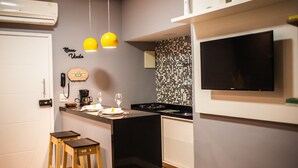 Una cocina compacta con estufa negra, armarios blancos, una pequeña isla, dos taburetes de madera, una cafetera y un televisor montado en la pared.
