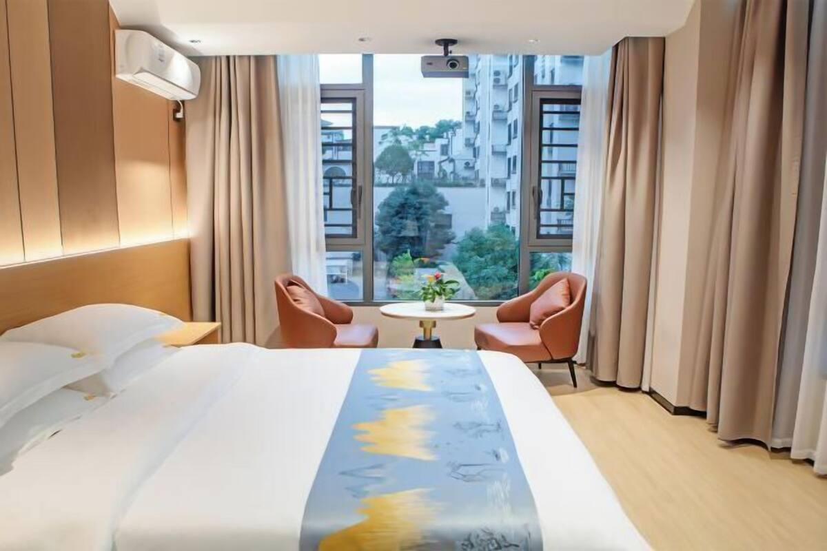 Hotellihuone, jossa on sänky, kaksi tuolia, pieni pöytä ja ikkuna, josta on näkymä rakennuksiin ja puihin.