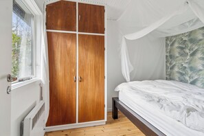 A bedroom with a wooden wardrobe, a bed with white bedding, and a window with a view of greenery.