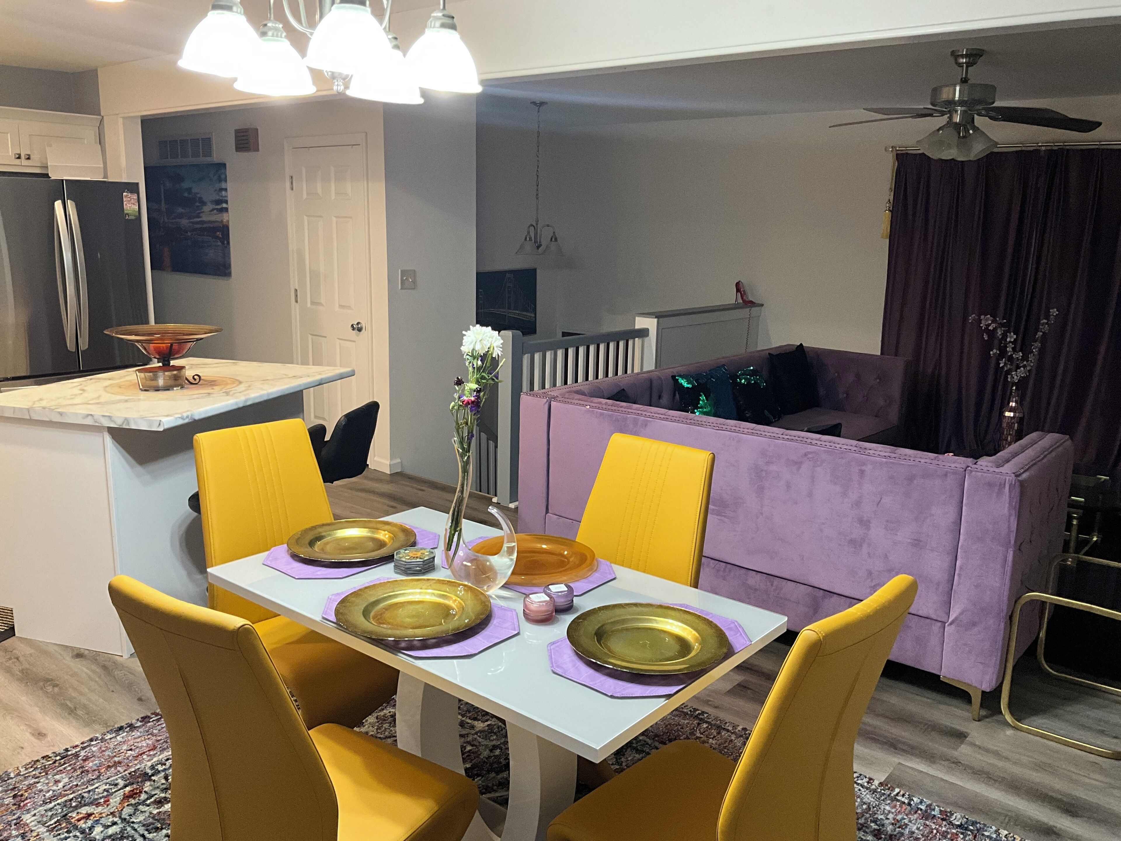 A modern kitchen with a dining area, featuring a white table with yellow chairs, a purple sofa, and a ceiling fan.
