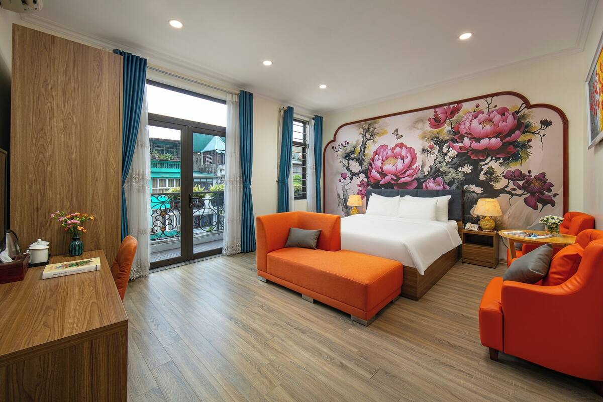 Une chambre d’hôtel avec un lit, un canapé orange, un bureau en bois et une grande fresque murale à motifs floraux.