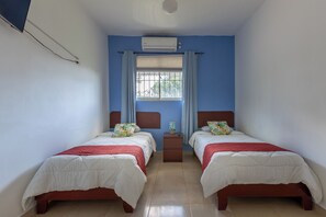 A bedroom with two beds, a window with curtains, an air conditioner, and a small bedside table.