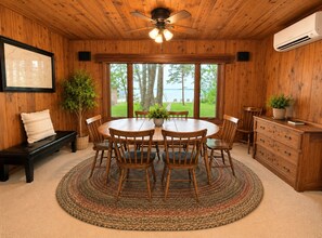 Ein Holzinterieur mit einem runden Tisch, Stühlen, einer Bank, einem Deckenventilator und Blick ins Freie.