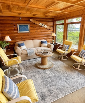 A cozy cabin interior with a wooden ceiling, a sofa, chairs, a cat tree, and large windows.
