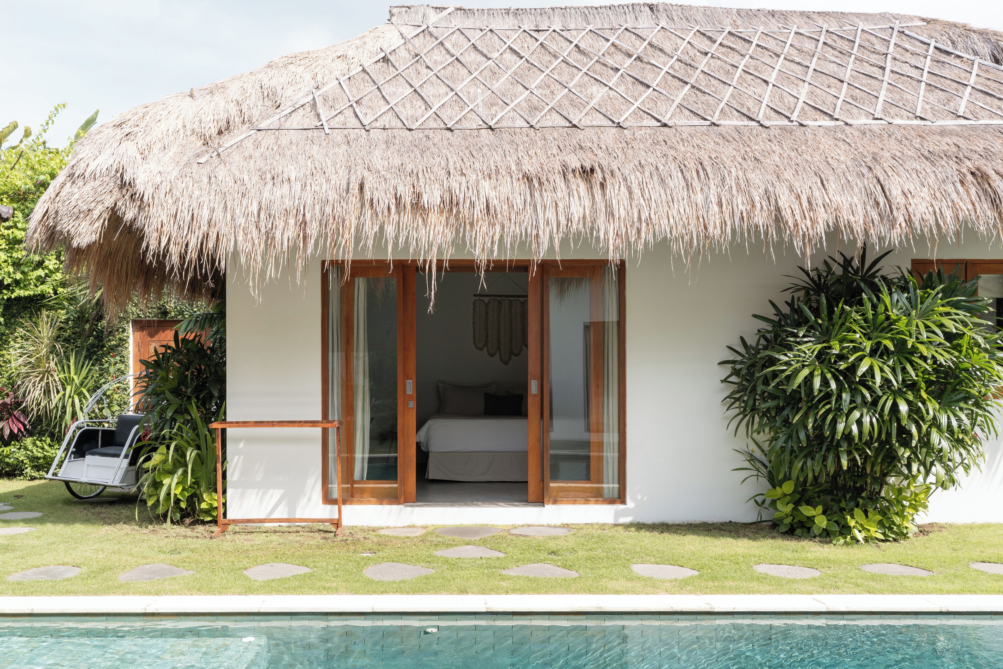 A thatched-roof bungalow with a pool, a bed, and a large plant.