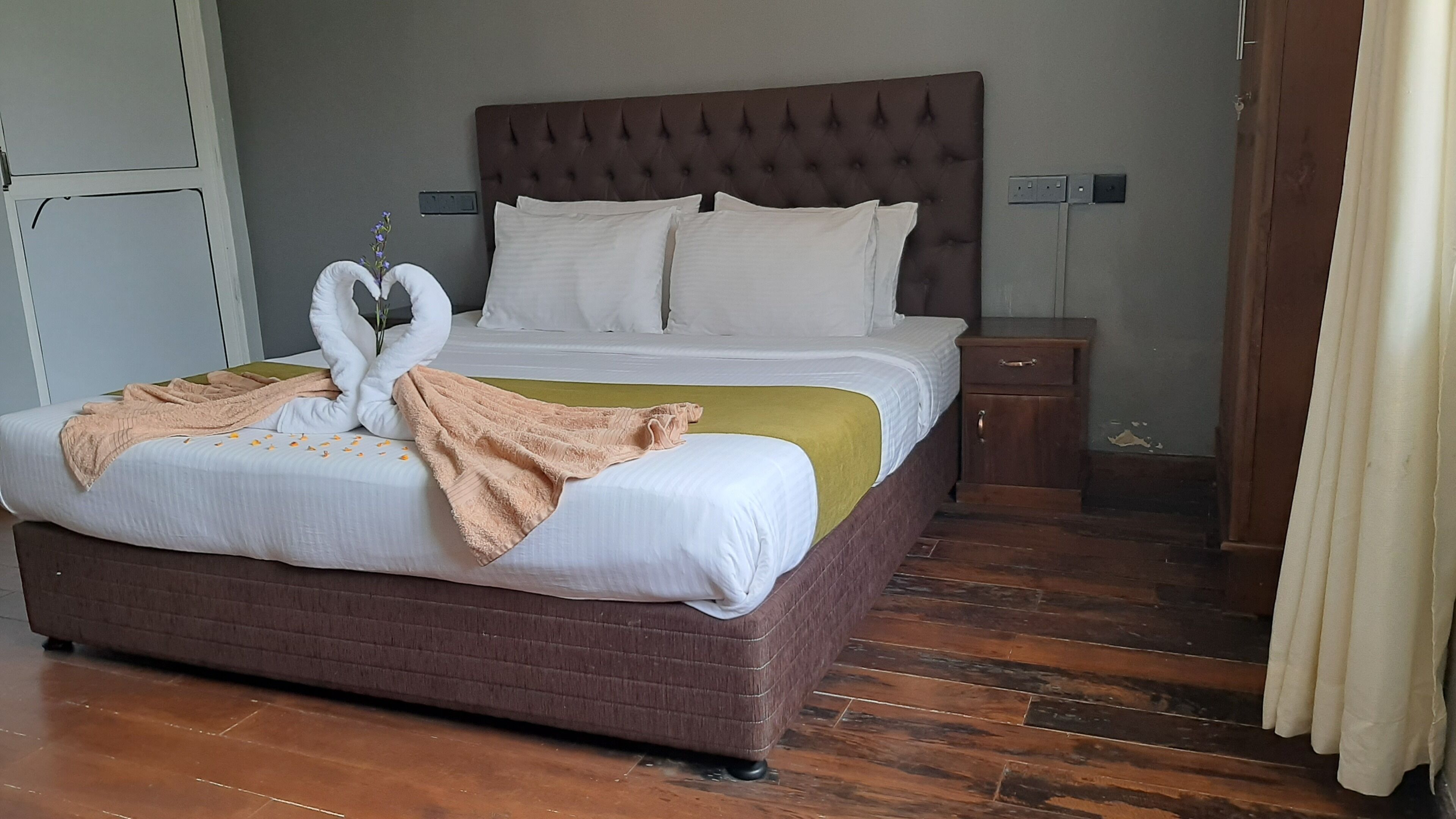 A neatly made bed with a brown headboard and white bedding, a wooden nightstand with a drawer, and a wooden floor.