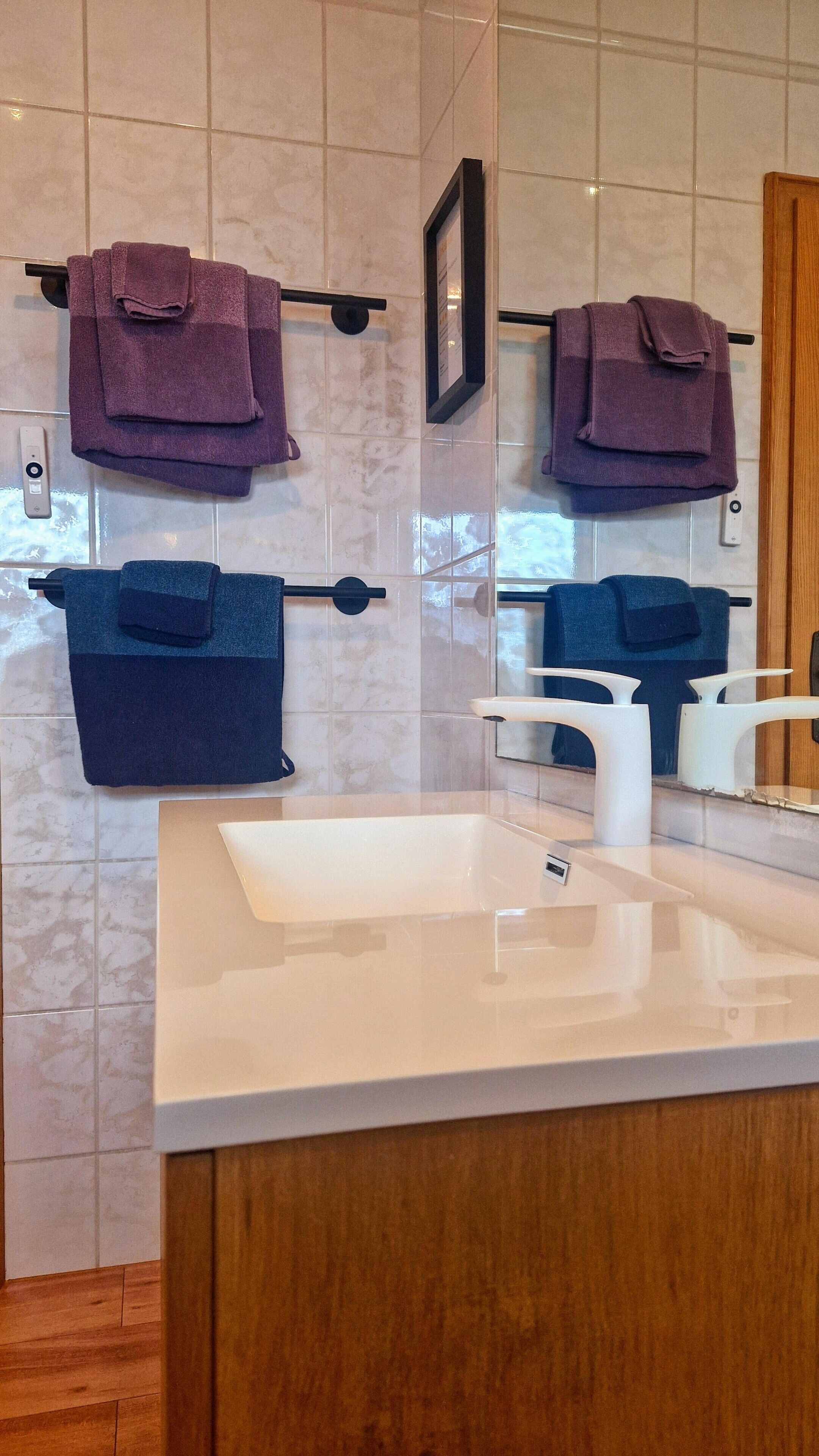 Ein Badezimmer mit einem weißen Waschbecken, zwei Handtuchhaltern mit lilafarbenen Handtüchern, einem Spiegel und einem gerahmten Bild an der Wand.