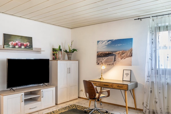 A room with a TV on a wooden stand, a desk with a chair, a potted plant, and a framed picture on the wall.