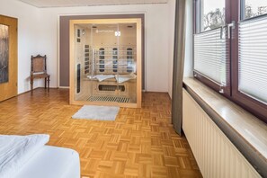 Une pièce dotée d’un parquet en bois, d’un lit, d’une fenêtre avec des stores et d’un sauna visible à travers une porte en verre.