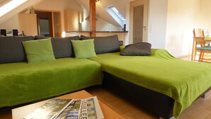 Ein Loft-Zimmer mit einem grünen Sofa, einem Holzesstisch und einer Küche im Hintergrund.