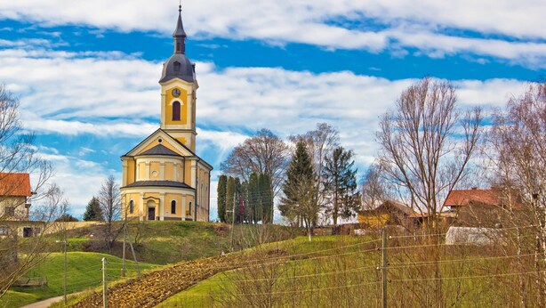 A church with a prominent bell tower, surrounded by a hilly landscape and sparse trees.