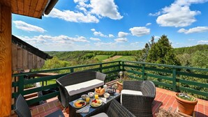 A balcony with wicker furniture, a table set with breakfast, and a scenic view of green hills and trees. - Hunting Lodge Hills Retreat, Ribnjačka, Croatia (Ribnjačka)