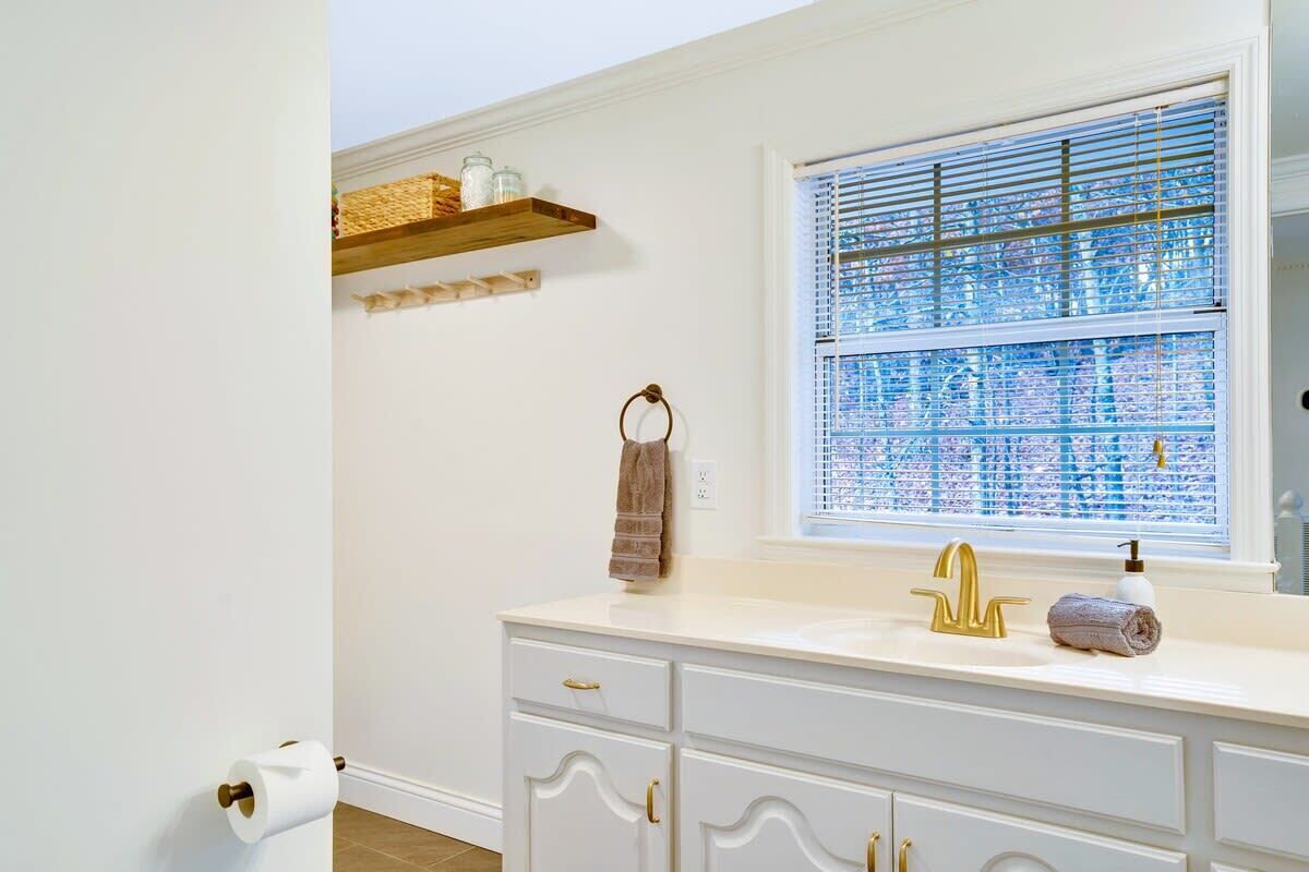 A bathroom with a white vanity, a gold faucet, a window with blinds, and a towel rack.