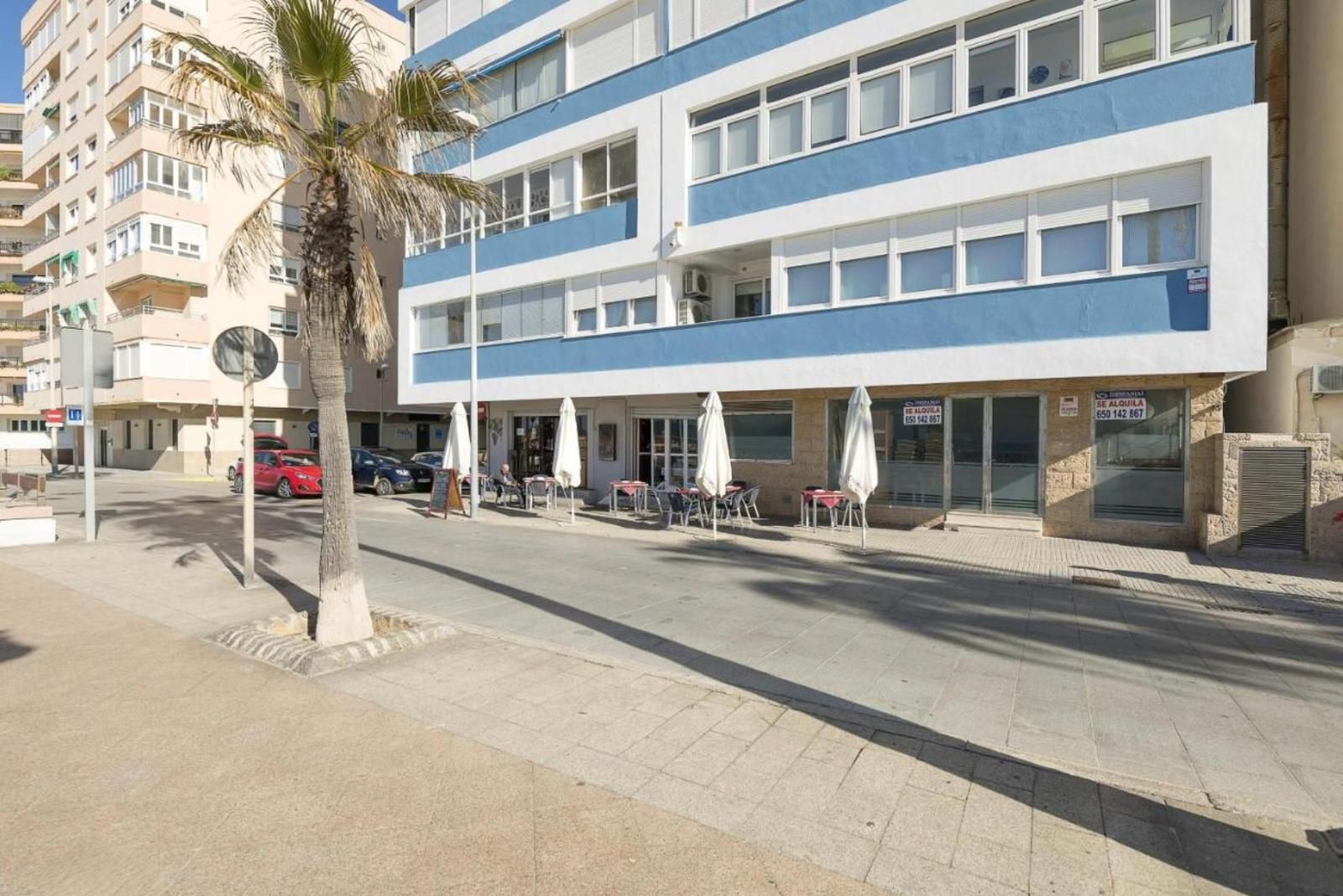 Ein mehrgeschossiges Hotel mit blau-weißer Fassade, einer Sitzgelegenheit im Freien mit Sonnenschirmen und einer Palme.