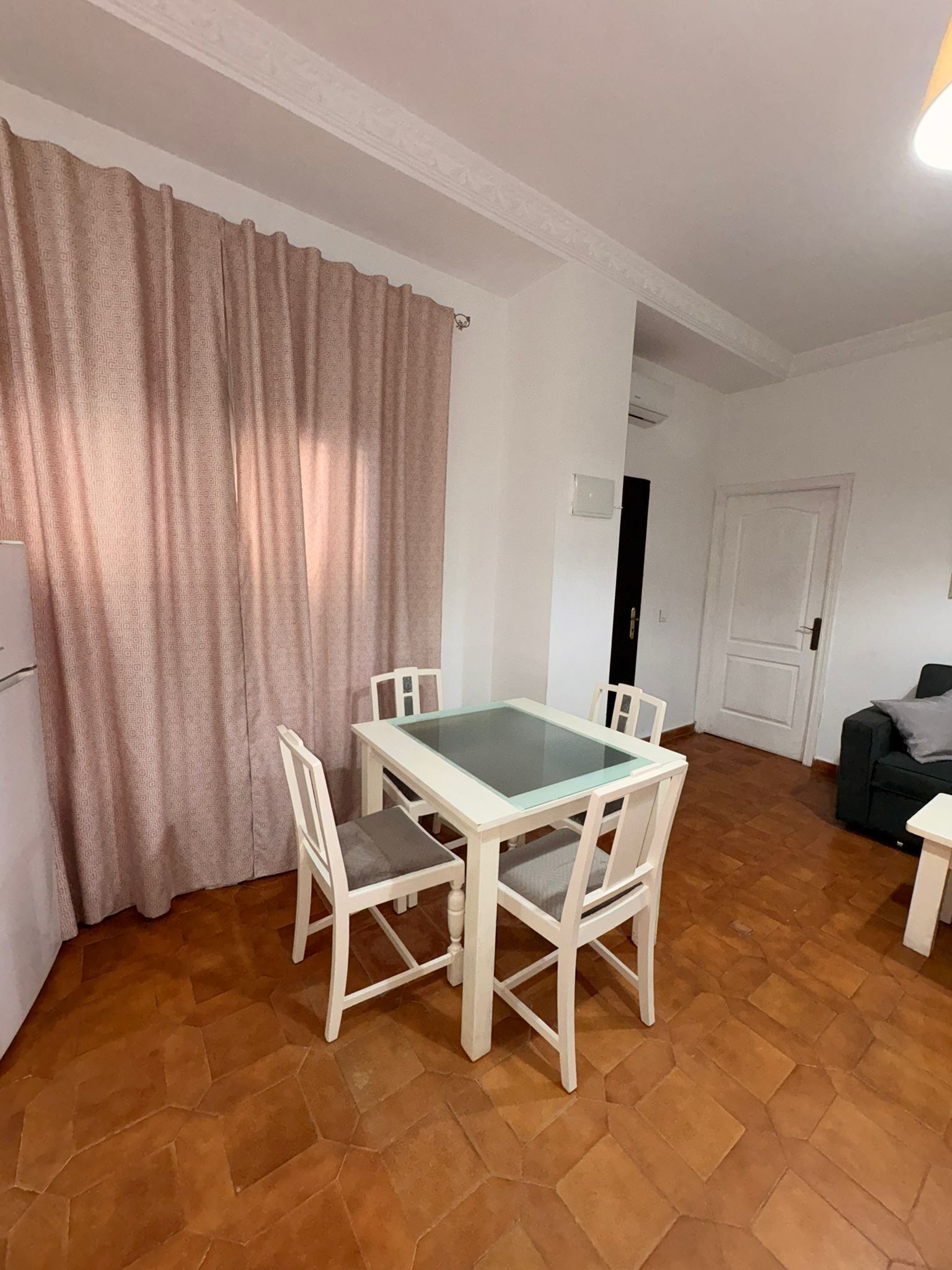 Een eetgedeelte met een tafel met glazen blad en witte stoelen, een keuken met een koelkast, en een woonruimte met een bank en een deur.