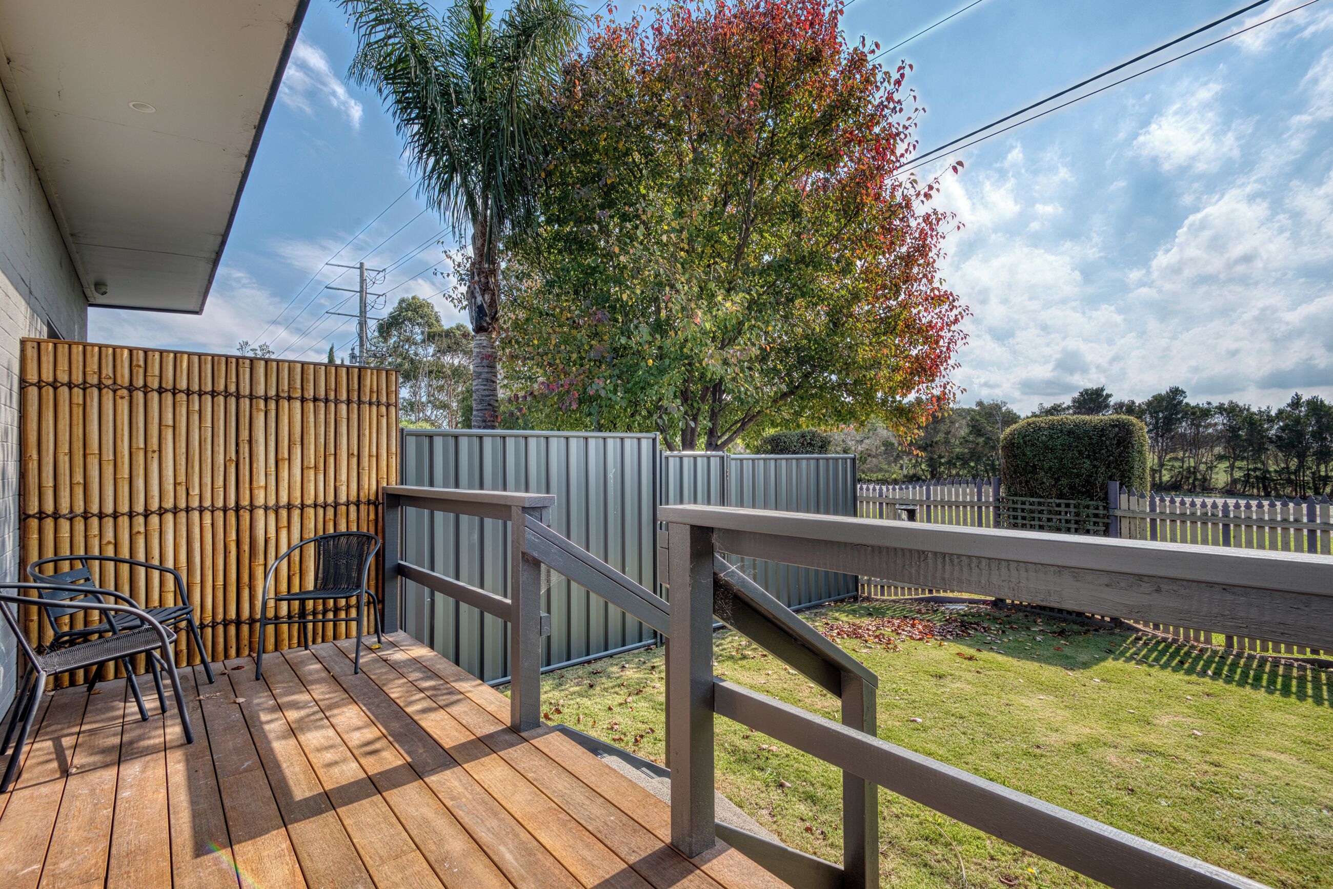 Une terrasse en bois avec des rampes en métal, une clôture en bambou et une vue sur un jardin avec des arbres et une pelouse.