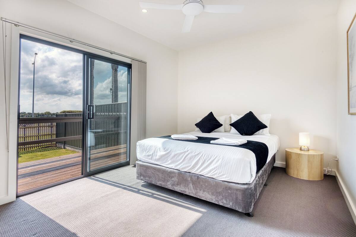 A modern bedroom with a large bed, a small bedside table with a lamp, and a sliding glass door leading to an outdoor area.