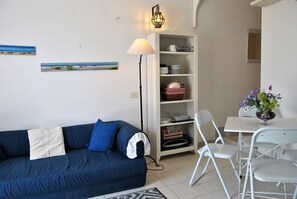 En stue med en blå sofa, et hvidt bord med stole, en gulvlampe og et hvidt reolsystem med tallerkener og en kurv.