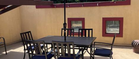 Un patio exterior con una mesa, sillas, un paraguas azul y una ventana con marco rojo.