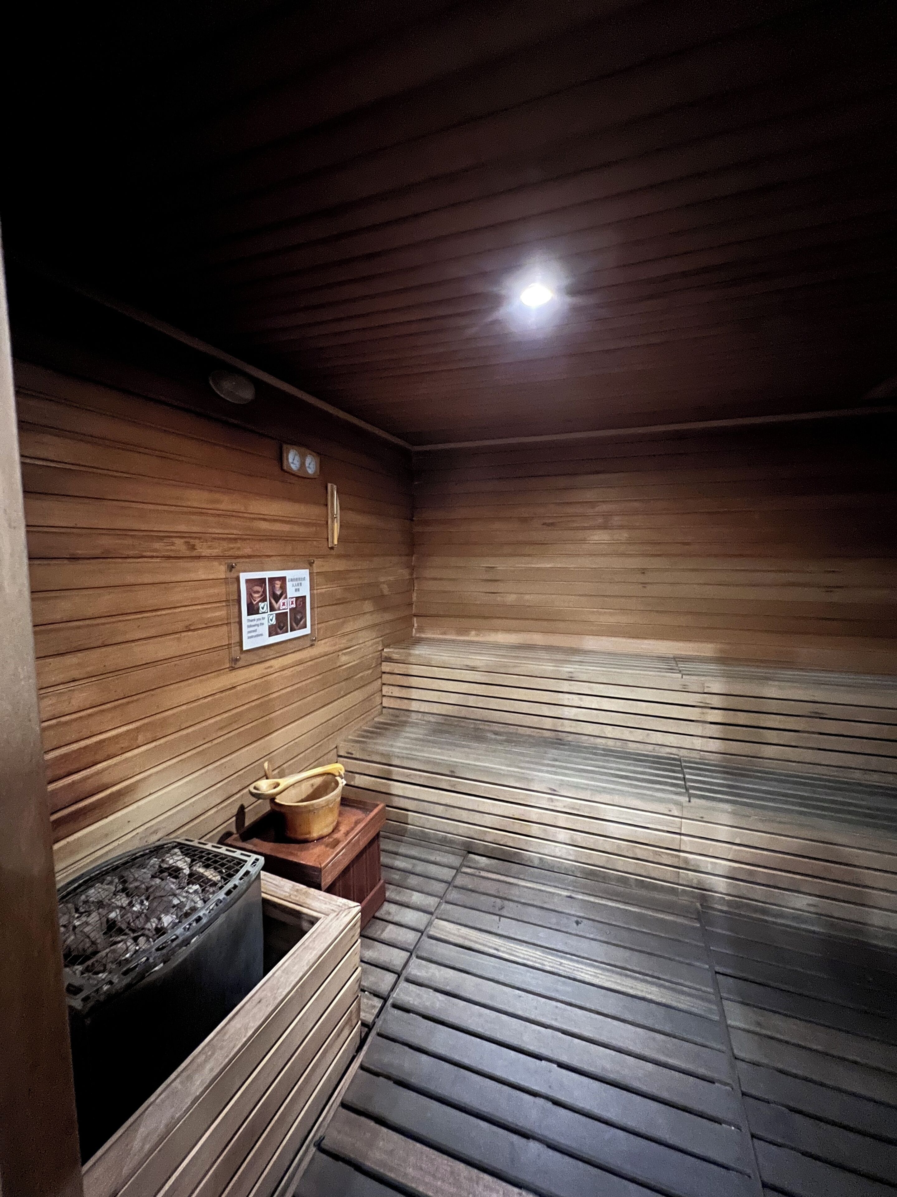 A sauna room with wooden walls and a sloped ceiling. There is a bench and a bucket on the floor.